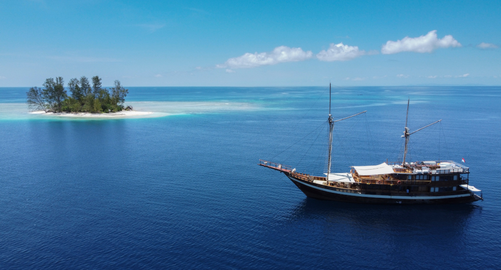 our phinisi cruise ship Coralia at sea next to a tiny tropical island in Raja Ampat