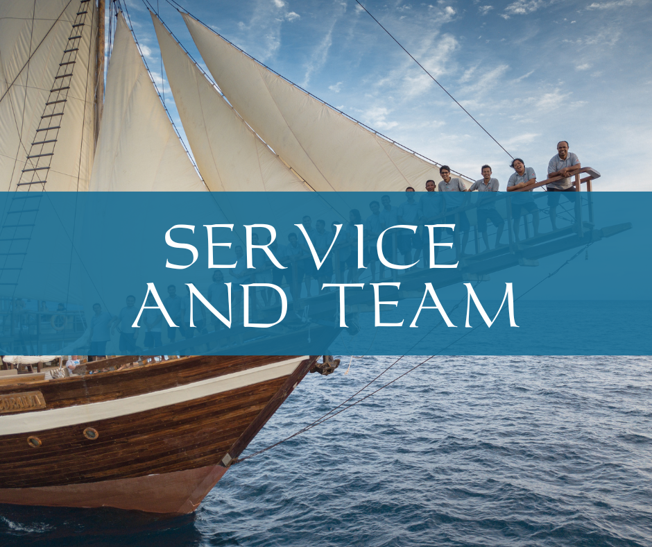 Crew members standing on the bow sprit of Coralia liveaboard under full sails in Indonesia.